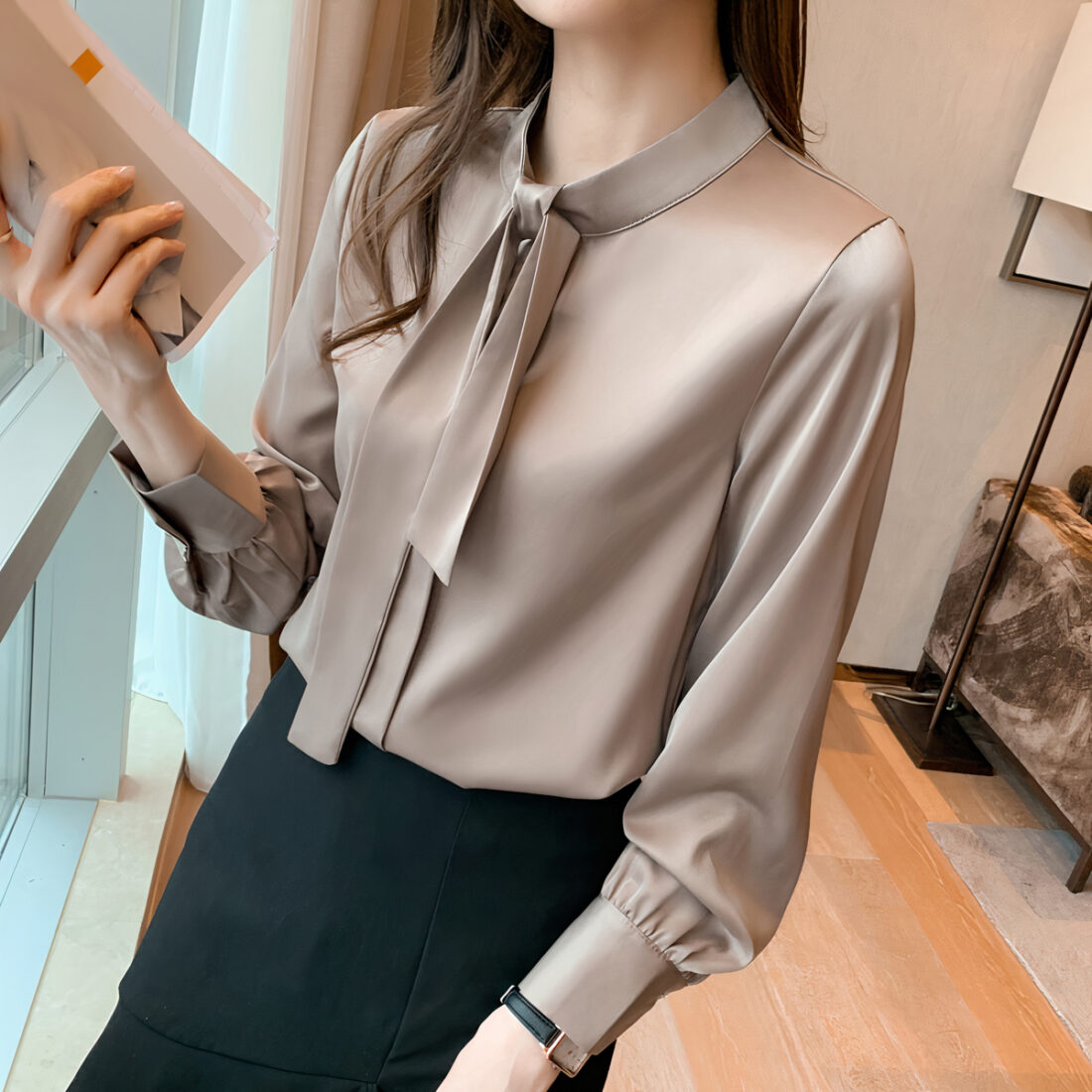 Femme portant une blouse en satin beige avec un noeud autour du cou. Cette femme tient un journal dans la main droite.