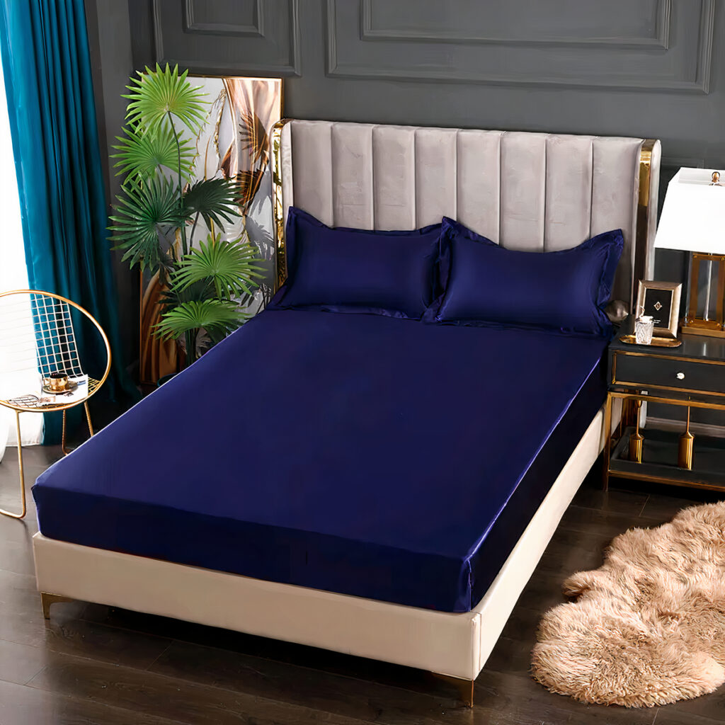 Drap housse en satin bleu marine sur lit double avec coussins assortis. Tête de lit grise, tableau style fourrure rose en bas à droite du lit. Table de chevet avec lampe à droite du lit. Plante verte et tableau à gauche, rideau turquoise à gauche.