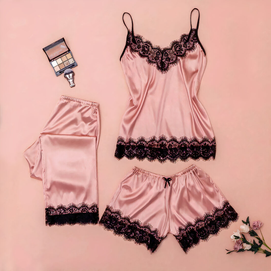 Pyjama 3 pièces rose composé d'un débardeur, un short et un pantalon en satin avec de la dentelle. Les trois vêtements sont posés à plat sur un fond rose avec une palette de maquillage à côté.