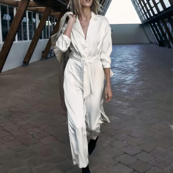 Femme portant une combinaison pantalon en satin blanc. La femme marche dans un espace industriel en tenant un manteau par dessus son épaule.