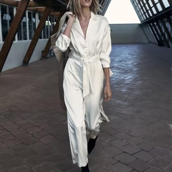 Femme portant une combinaison pantalon en satin blanc. La femme marche dans un espace industriel en tenant un manteau par dessus son épaule.