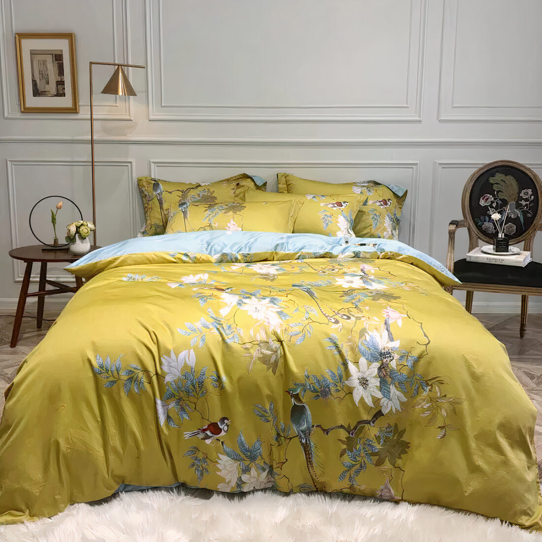 Lit avec des draps jaunes aux motifs de fleurs et d'oiseaux dans une chambre avec du parquet, des fleurs blanches et un miroir rond au mur