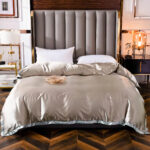 Lit avec une couette en satin gris dans une chambre avec du parquet et une tête de lit grise matelassée, des tables de chevet de chaque côté avec des lampes contemporaines, un plat rond marron sur le lit.
