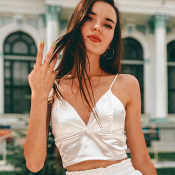 Crop top en satin blanc à bretelles porté par une femme brune aux cheveux détachés debout dans la rue, on ne la voit qu'au-dessus de la taille, main droite relevé formant la lettre V avec ses doigts.