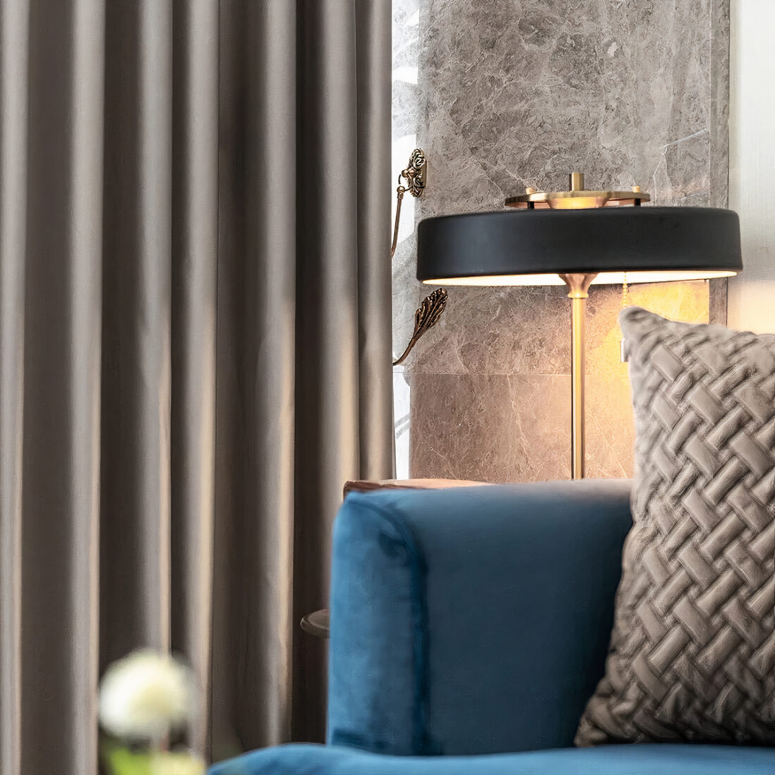 Bord de canapé bleu avec un coussin dessus. Derrière se trouve une lmape noire sur pied avec un rideau gris.