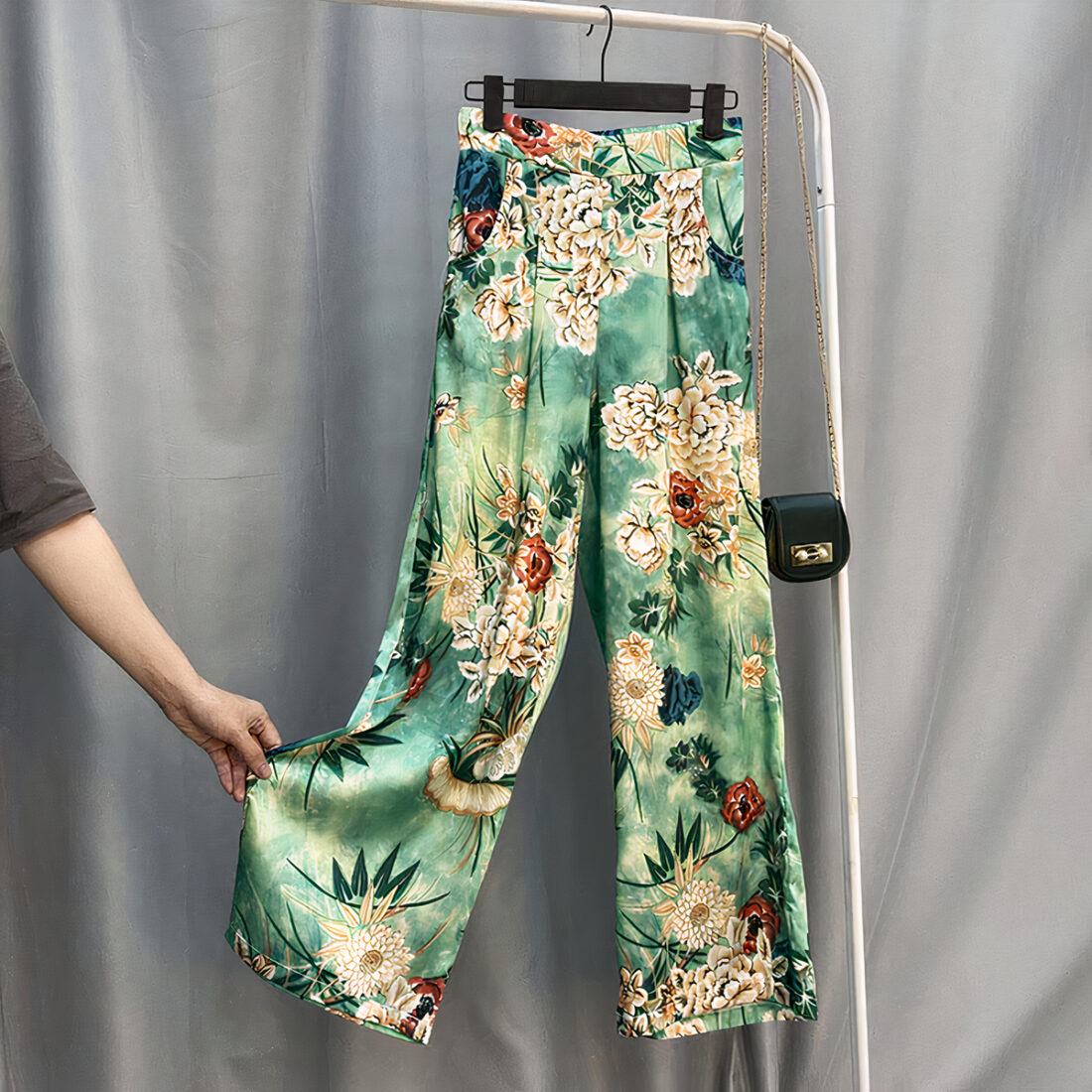 Pantalon en satin vert avec motif à fleurs posé sur un ceintre. Une main soulève le bas de la jambe droite