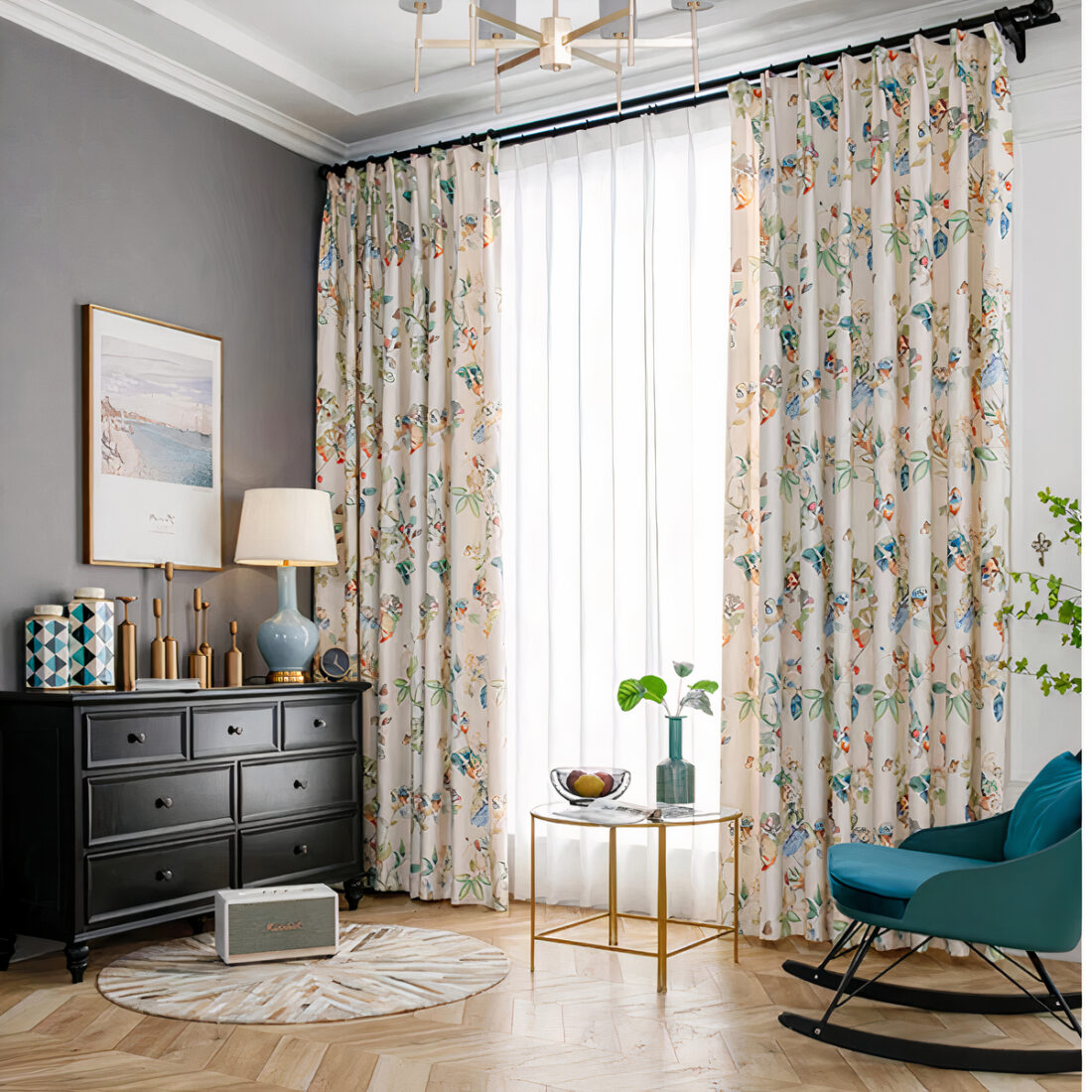 Rideau beige avec des motifs d'oiseaux et de fleurs dans une pièce avec mur gris sur la gauche, buffet en bois marron, des objets dessus dont une lampe, un tapis rond devant, une petite table devant la fenêtre et un rockingchair turquoise sur la droite.