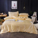 Photo d'un lit avec des draps de satin beige. 4 coussins assortis. un mur noir avec un cadre blanc. une table de chevet de chaque côté avec des objets dessus.