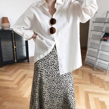 Femme debout dans une pièce avec une jupe léopard, un chemise blanche et des lunettes de soleil accrochées sur sa chemise. Sa main droite posée sur sa taille Derrière se trouve un bureau et une étagère grise. Le sol est marron clair.