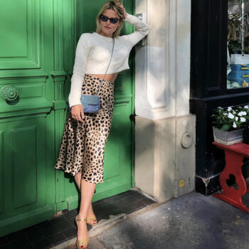 Femme en jupe léopard et haut blanc avec des lunettes de soleil se tenant dans la rue devant une porte verte