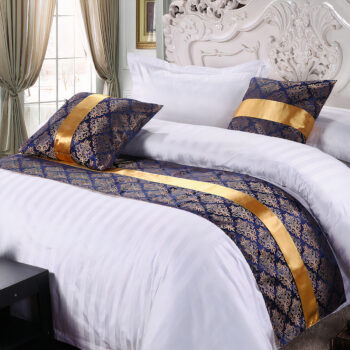Couvre lit en satin à motifs Jacquard dorés sur fond bleu. Il est posé sur un lit avec une literie blanche. Dans une chambre avec tête de lit vintage et fenêtre avec rideaux plissés beige.