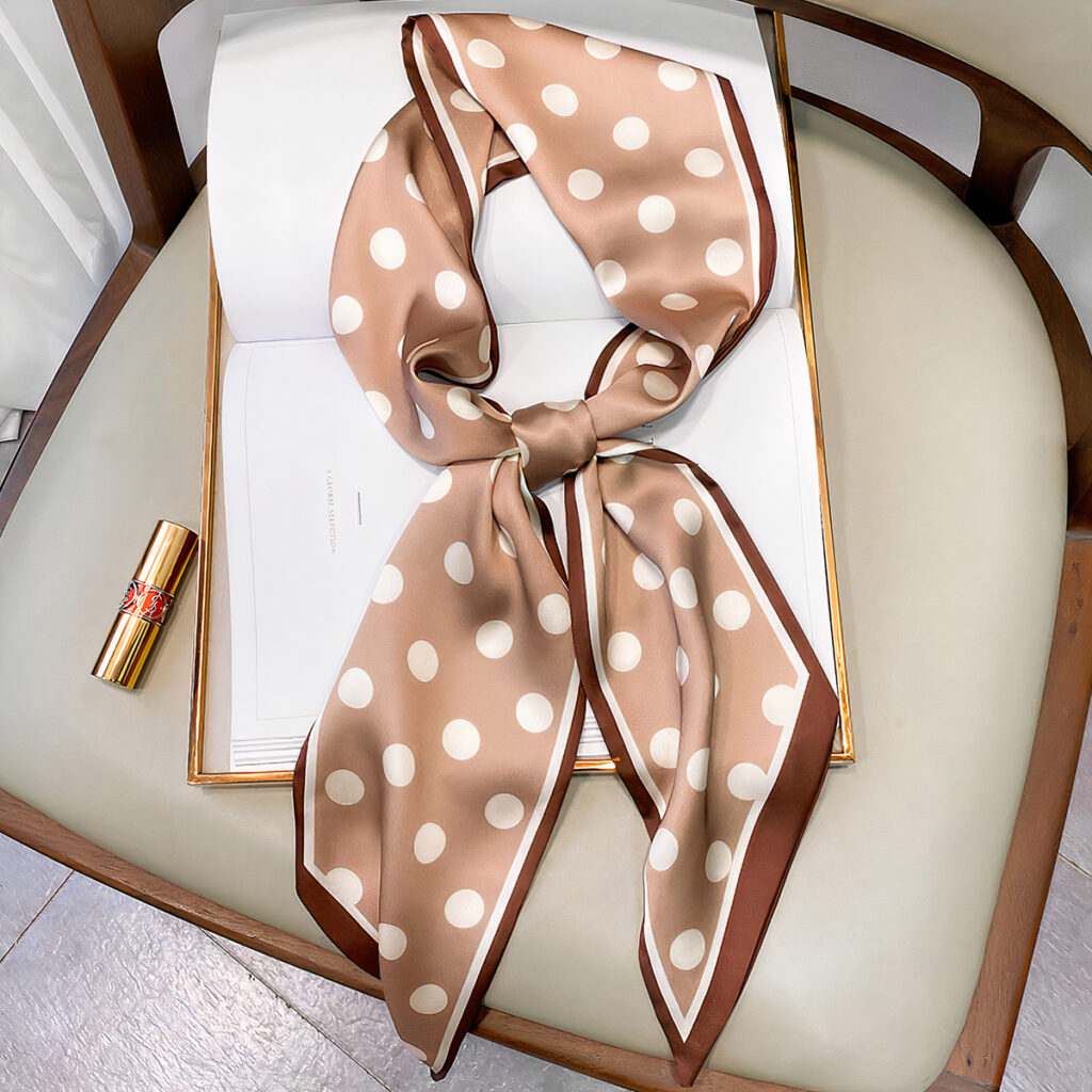 Foulard en satin beige à pois blanc, noué, posé sur une chaise avec un rouge à lèvres