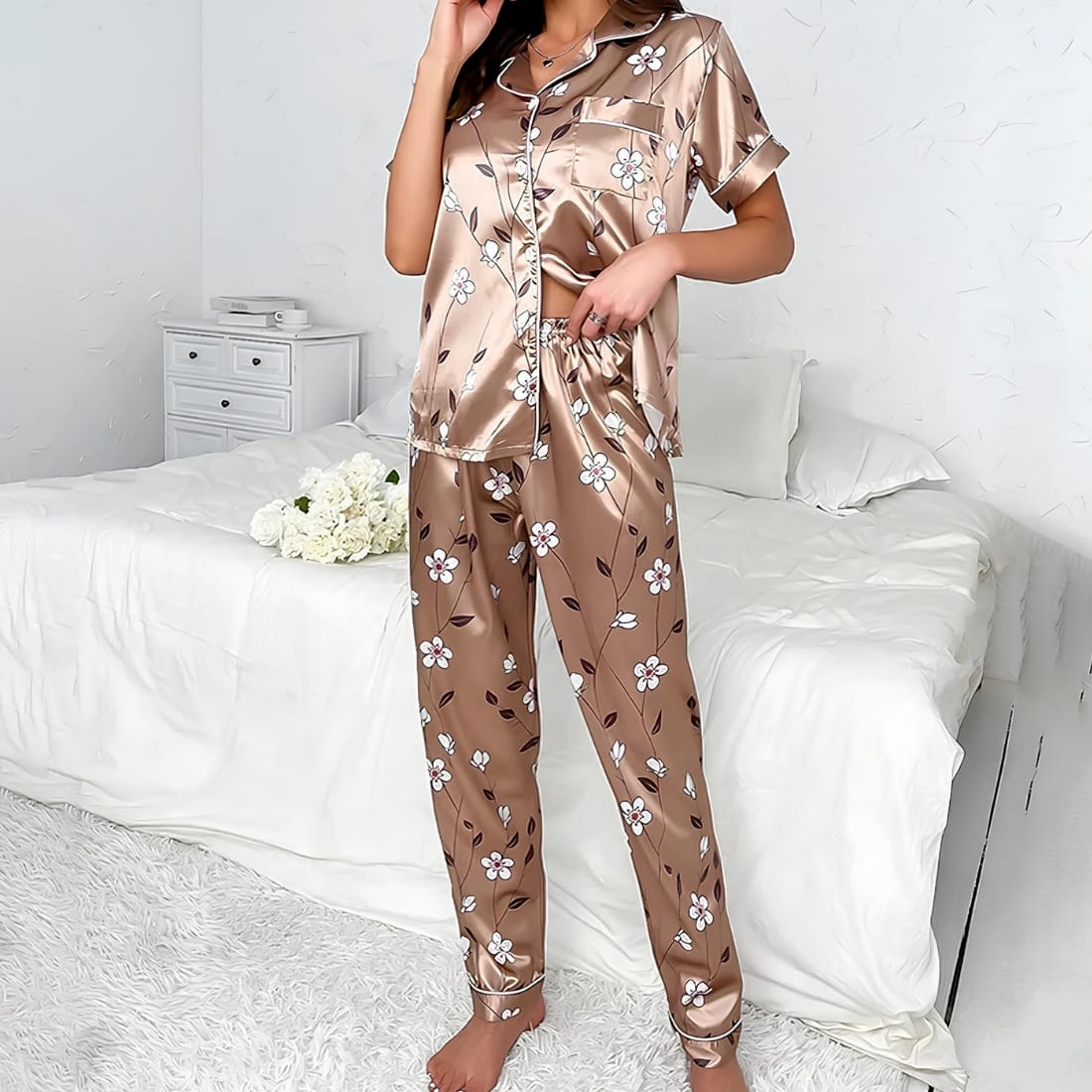 Ensemble de pyjama en satin beige pour femme. Le bas est un pantalon, le haut est une chemise à manches courtes. L'ensemble a des motifs fleuris. Porté par une femme devant un lit blanc.