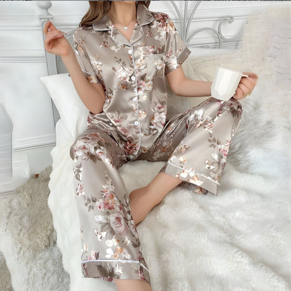 Ensemble de pyjama en satin beige pour femme. Le bas est un pantalon, le haut est une chemise à manches courtes. L'ensemble a des motifs fleuris. Porté par une femme devant un lit blanc.