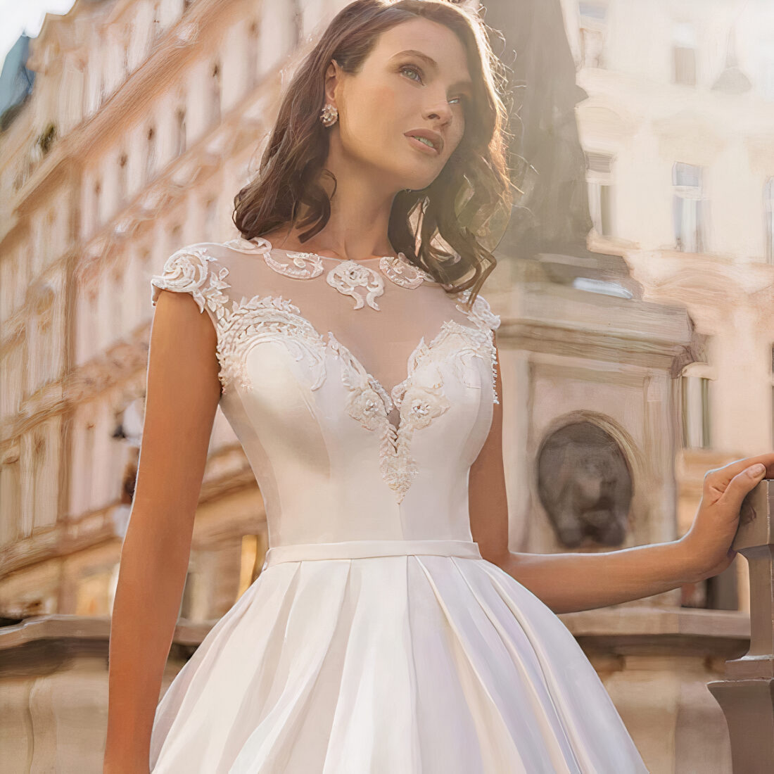 Robe de mariée évasée en satin portée par une femme dans la rue avec les cheveux jusqu'aux épaules, sa main gauche posée sur un muret.