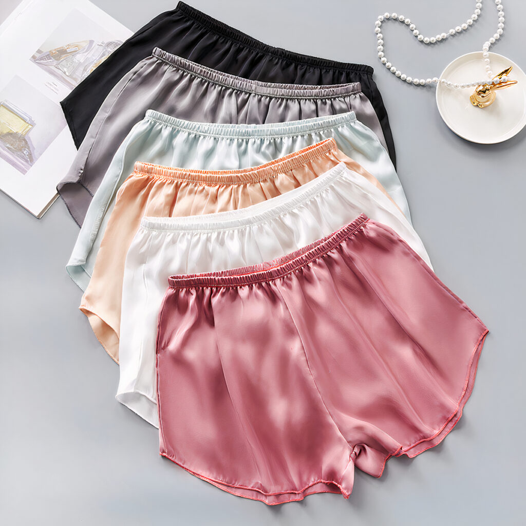 Ensemble de six shorts en satin, taille élastique. Ils sont bien présentés, l'un par-dessus l'autre comme dans une boutique, et il y a un porte bijous et un collier en perles à côté.