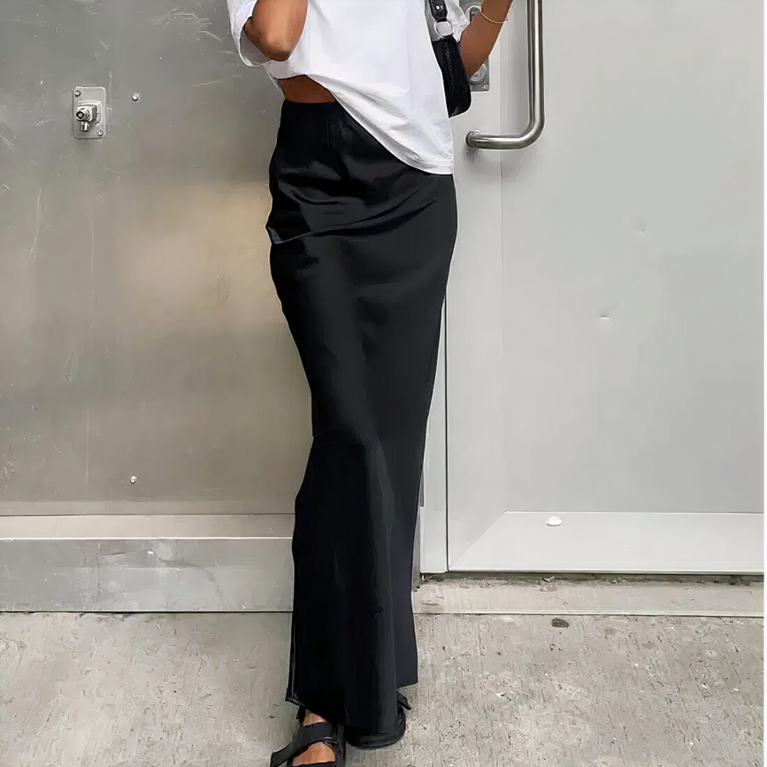 Une femme porte une jupe longue de couleur noire en satin, de coupe slim. Elle a un haut blanc, un sac noir sur l'épaule gauche et des sandales noires aux pieds. Elle debout sur un sol en béton gris et derrière elle se trouve un mur de couleur aluminium et une porte grise.