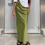 Robe longue verte en satin avec une coupe slim, portée par une femme avec des sandales noires, un haut blanc et un sac à main noir sur l'épaule gauche. Elle est debout sur un sol en béton, devant une porte grise.