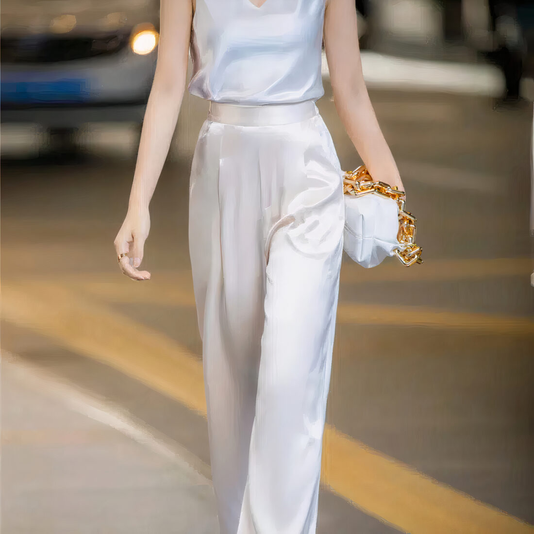 Femme devout portant un pantalon beige en satin a jambes larges et un haut blanc sans manche satiné. Elle a un sac à main blanc avec chaîne dorée dans sa main gauche. On voit la rue derrière elle.