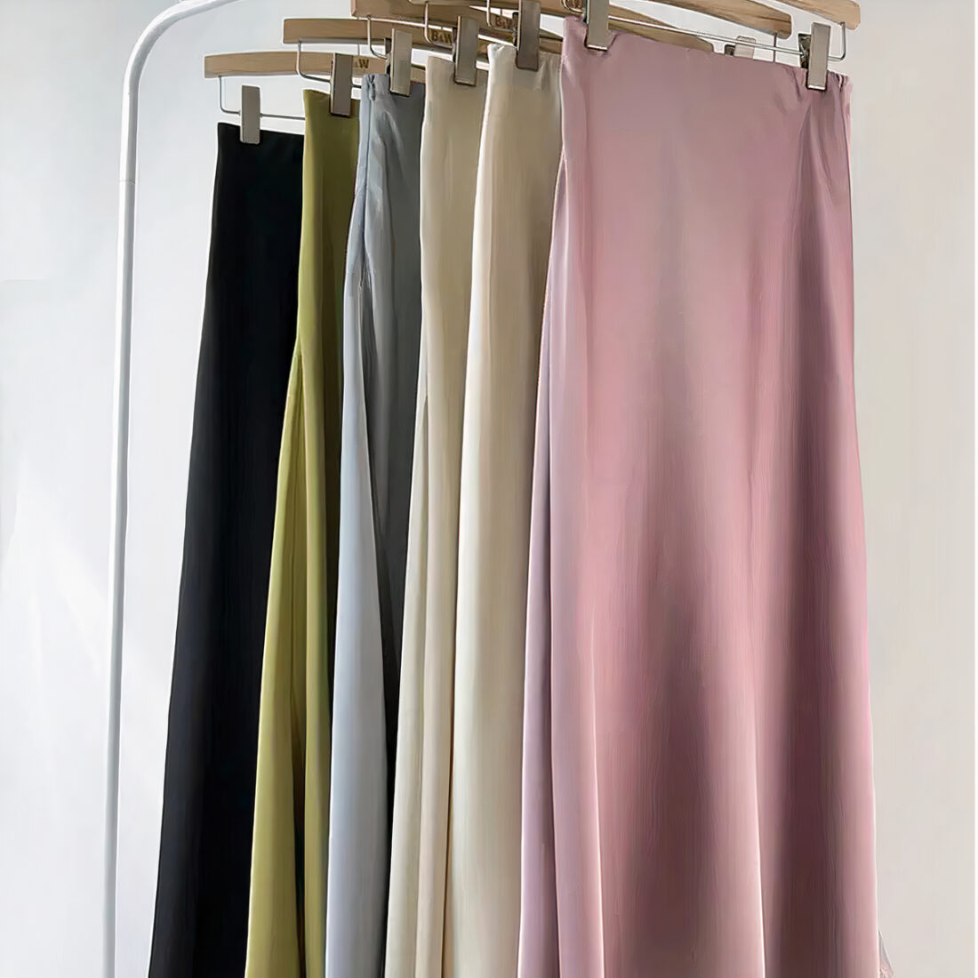 6 jupes les unes derrière les autres accrochées chacune à un cintre à pinces. Les cintres en bois sont accrochés à un support en métal blanc. Les jupes sont noire, verte, grise, 2 beiges et une rose en partant de la dernière à celle de devant.