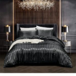 Lit 2 places avec literie en satin à rayures ton sur ton noires. Tête de lit noire. 2 coussins assortis et 2 coussins blancs derrière. un linge blanc est posé sur le milieu du lit. 2 tables de chevet de chaque côté avec lampe de chevet allumées identiques. Un lustre allumé en forme de branchage au dessus du lit. Mur marron sur la largeur du lit et extérieur gris.