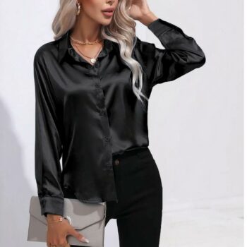 Femme blonde cheveux détachés ondulés portant une chemise noire en satin boutonnée, une moitié dans son pantalon noir. Dans sa main droite une pochette argentée, sa main gauche à son visage.