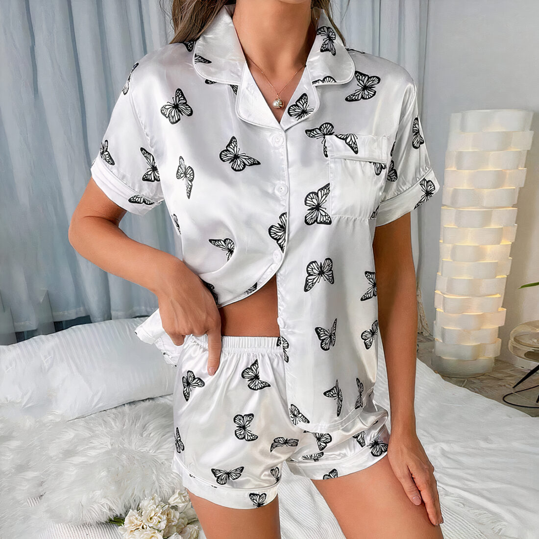 Femme portant une pyjama court avec des motifs de papillons. Elle est debout sur un lit, derrière elle se trouve une lampe blanche haute. Son bras gauche est le long de son corps, la main droite glissé dans le bord du short laissant apparaître un peu son ventre.