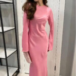 Robe longue à manches longues évasées en satin de couleur rose portée par une femme aux cheveux longs détachés. Elle a dans sa main droite une pochette beige. A gauche une étagère noire et blanche, derrière la femme une porte blanche, les murs sont gris.