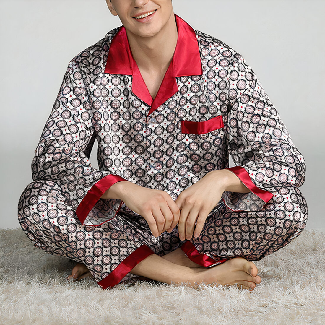 Homme pieds nus, assis en tailleur sur un tapis beige porte un pyjama en satin à motifs géométriques gris avec les bordures rouges. Ses coudes sont posés sur ses genoux.