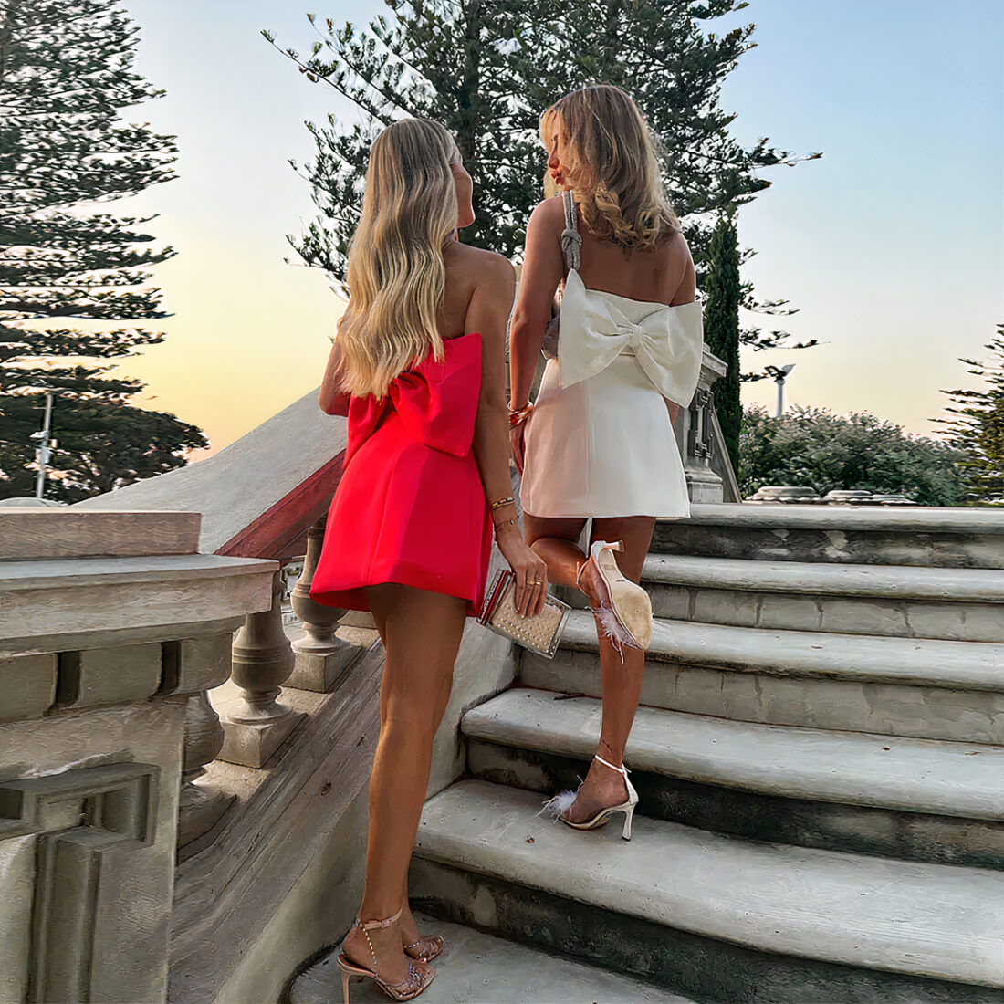 deux femmes blondes, cheveux détachés montant des escalier en pierre. Elles sont de dos, celle de gauche porte une robe courte bustier rouge en satin avec un gros nœud dans le bas du dos, des escarpins ouverts aux pieds, une pochette brillante dans sa main droite. La femme de droite porte une robe courte bustier blanche en satin avec un gros noeud dans le bas du dos, des escarpins ouverts aux pieds et un sac à min gris sur l'épaule gauche. Des sapins au fond.