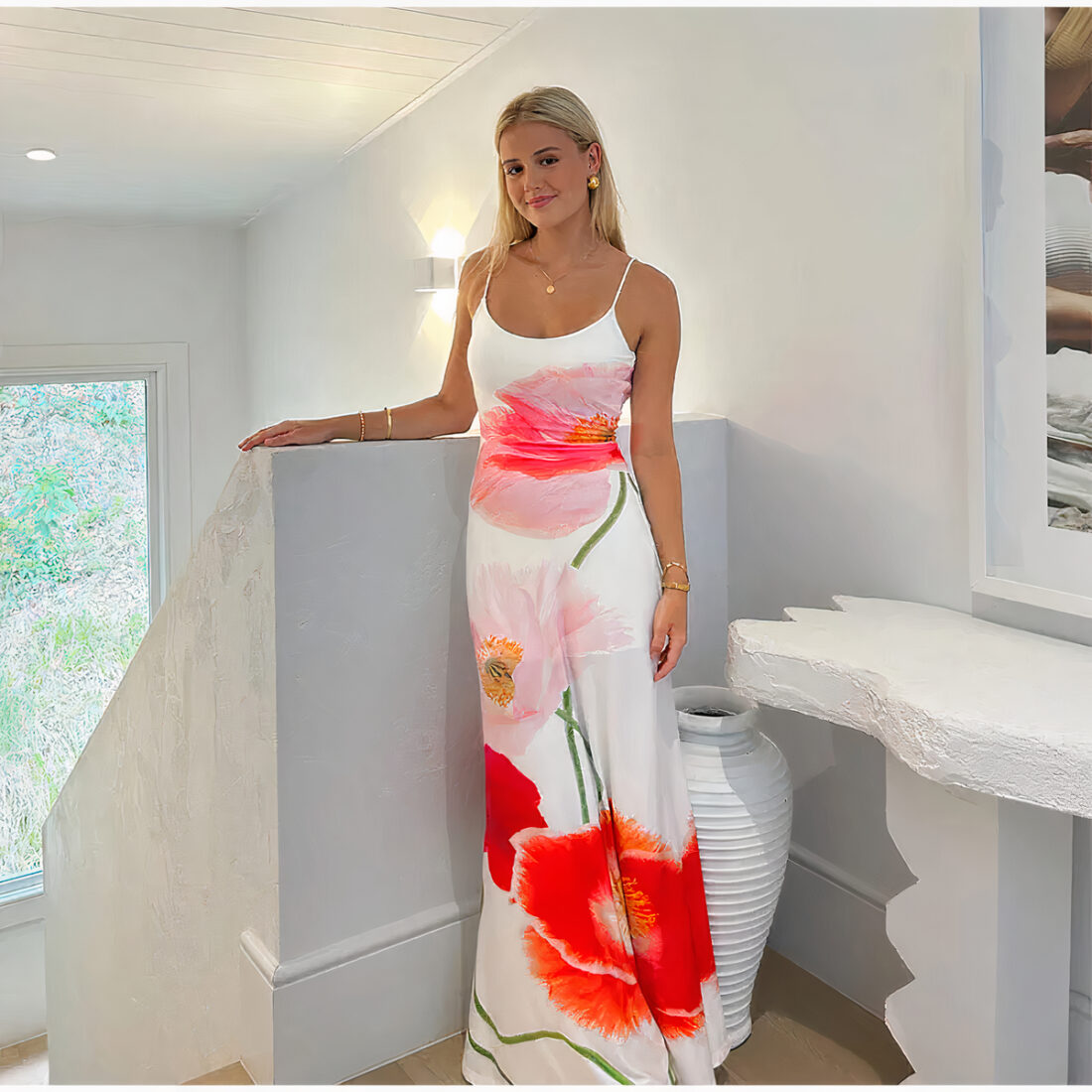 Femme blonde cheveux détachés, debout porte une robe longue à bretelle blanche avec des motifs de fleurs rouges et roses. Sa main droite est posé sur le muret de l'escalier qui descend à sa gauche. Un muret blanc est à sa gauche avec au-dessus un cadre. Une fenêtre au fond à gauche et l'éclairage derrière elle.