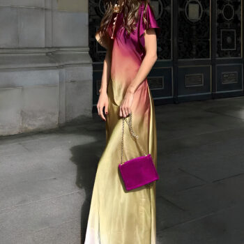 Femme de profil dans la rue avec cheveux longs portant une robe longue ample à manches courte avec un dégradé de couleurs du haut violet au bas jaune. Elle tient un sac à main violet avec une chaine dans ses mains. mur en pierre derrière avec grande grille en fer vert et noir.