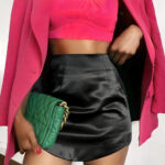 Femme métisse debout que l'on voit de la poitrine aux cuisses. Elle porte une mini jupe en satin noire, un haut rose laissant apparaître son ventre, une veste de la même couleur que le haut et un sac à main vert avec une chaîne dorée dans la main droite. Sa main gauche est posée sur sa taille.