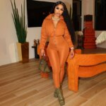 Femme debout métisse, cheveux noirs bouclés détachés, portant une combinaison en satin de couleur orange, la partie basse est slim comme un legging, la partie haute matelassée comme une doudoune. Elle a un sac à main marron dans sa main droite, la jambe droite légèrement devant la gauche. Derrière elle, un fauteuil orange, au fond un écran plat accroché au mur, une plante verte dans un vase doré et des valises orange au fond à droite empilées.