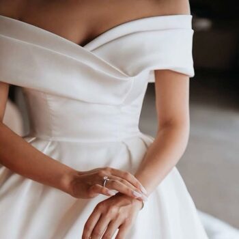 Femme portant une robe de mariée blanche avec épaules dénudées et cintrée à la taille. On la voit de son cou à son bassin. Elle son bracelet à son poignet gauche avec sa main droite. Elle a une bague à l'annulaire droit.